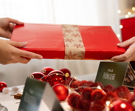 Zwei Hände übergeben ein rotes Geschenk mit einer Weihnachtsbanderole. Weihnachtsdeko ist im Hintergrund sichtbar.