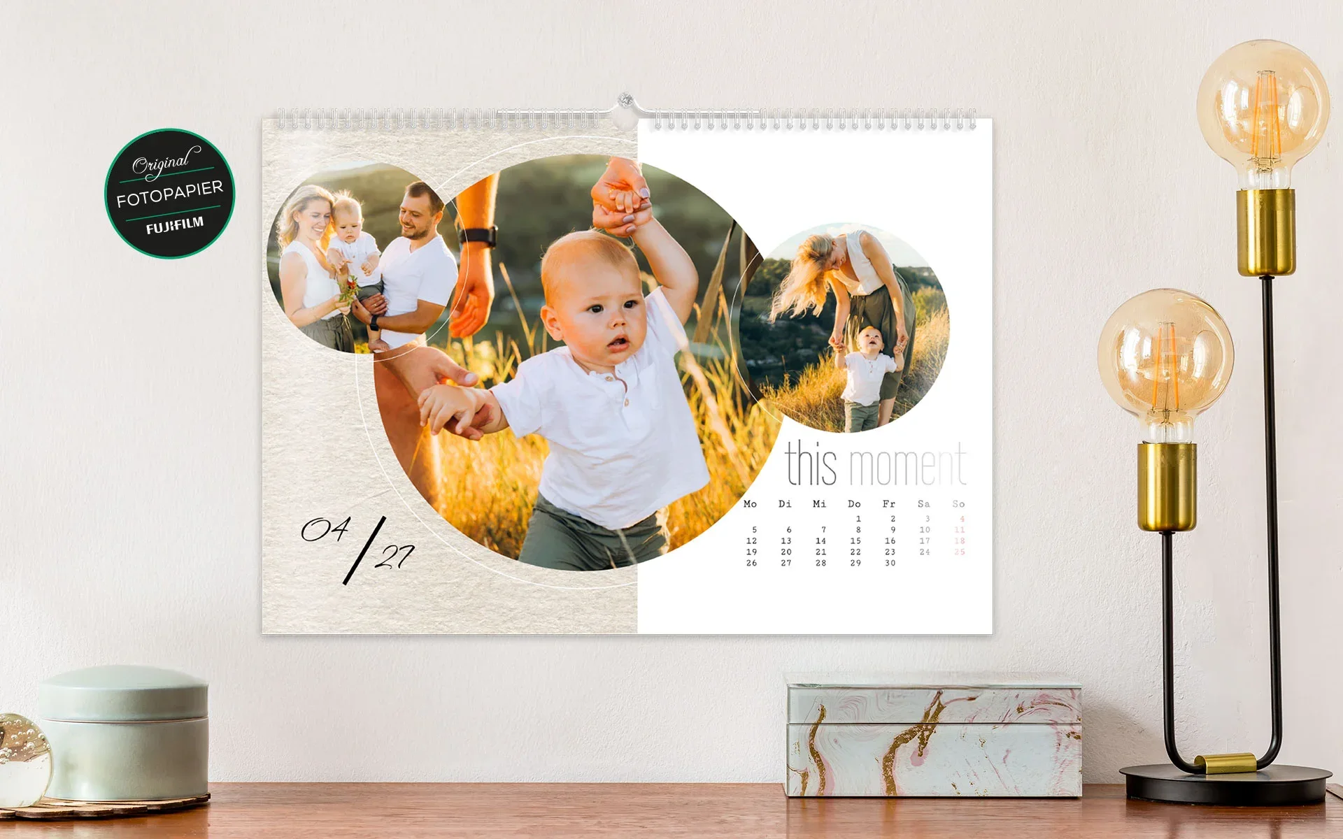 HappyFotokalender ca. A4 quer – aufgehängt Ein aufgehängter HappyFotokalender ca. A4 quer mit dem Text This Moment zeigt 3 Fotos einer Familie in kreisförmigen Rahmen.