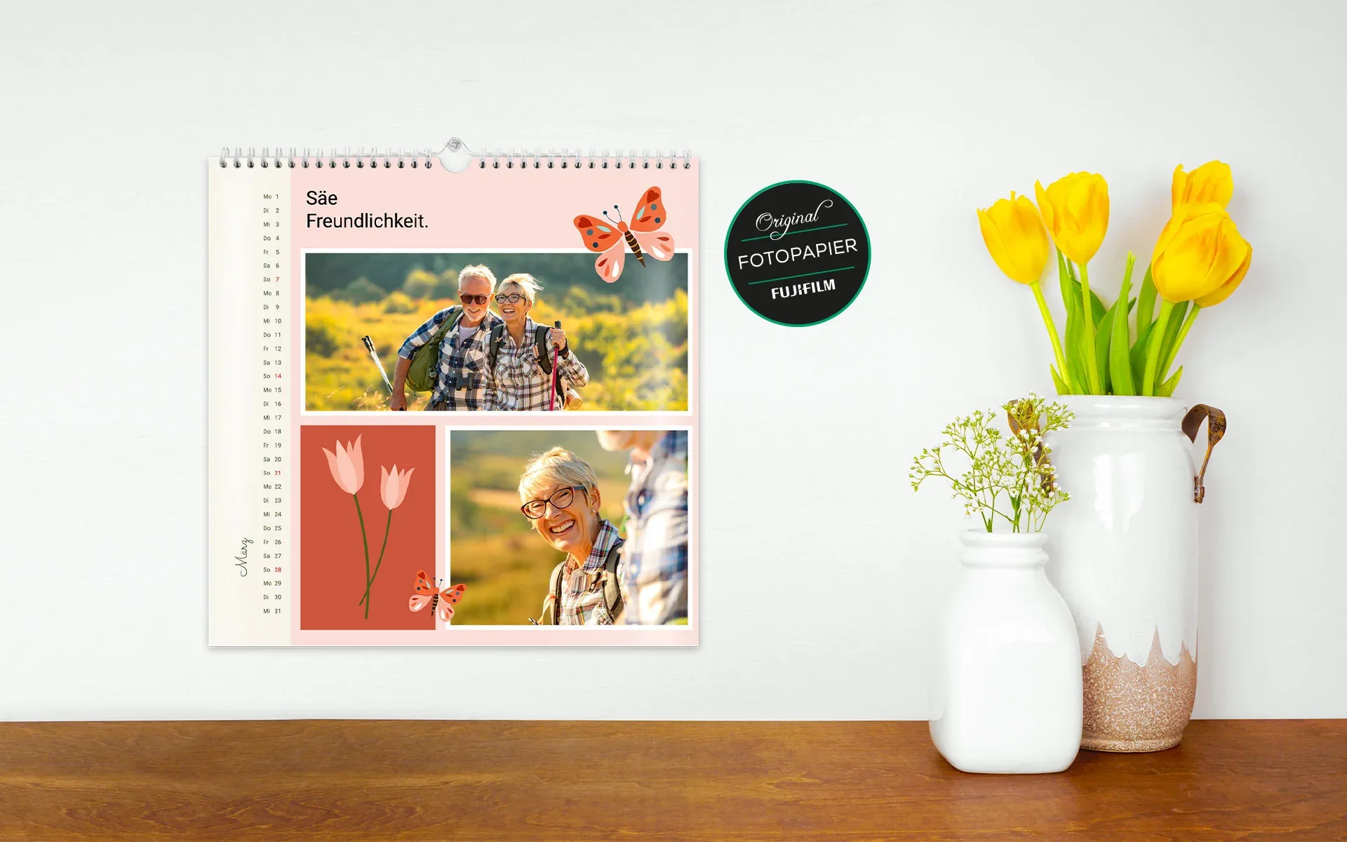 HappyFotokalender ca. 30 x 30 cm – aufgehängt Ein aufgehängter HappyFotokalender ca. 30 x 30 cm mit dem Text Säe Freundlichkeit zeigt 2 Fotos eines Paares vor rosa Hintergrund und Schmetterling Cliparts.