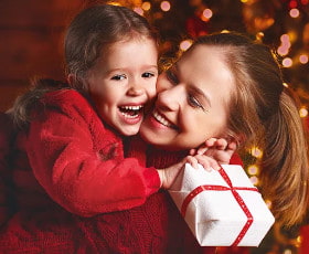 Eine Frau umarmt ein fröhliches Kind und hält ein Geschenk in den Händen, vor festlicher Weihnachtsdekoration.